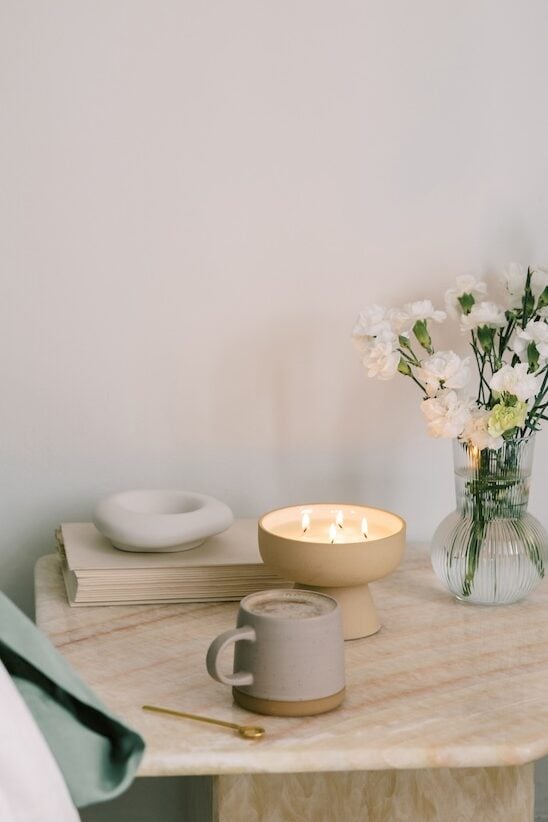 Bedside table with a lit candle, coffee mug, and flowers, creating a cozy morning environment.